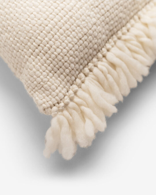 Detalhes das franjas de uma almofada quadrada feita a mão em tear com lã merino cor cru natural.